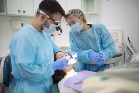 Dentist and her assistant working on young female patient in medical clinic with protective equipment,
